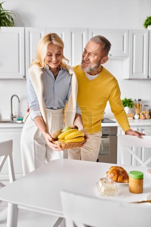 A mature loving couple in cozy homewear standing in a kitchen, holding bananas.