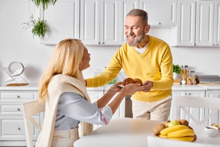 Ein reifer Mann und eine reife Frau in gemütlicher Kleidung stehen an einem Küchentisch und teilen einen zarten Moment, während sie zu Hause gemeinsam eine Mahlzeit zubereiten.