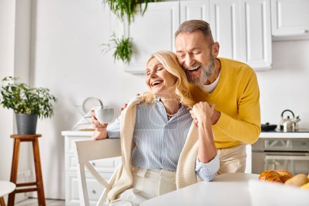 A mature loving couple, dressed in cozy homewear, share a moment of genuine laughter and joy in their kitchen at home.