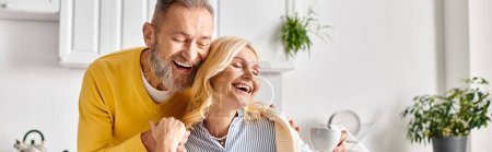 Ein reifer Mann und eine reife Frau in Hauskleidung teilen einen freudigen Moment, als sie gemeinsam in einer gemütlichen Küche zu Hause lachen.
