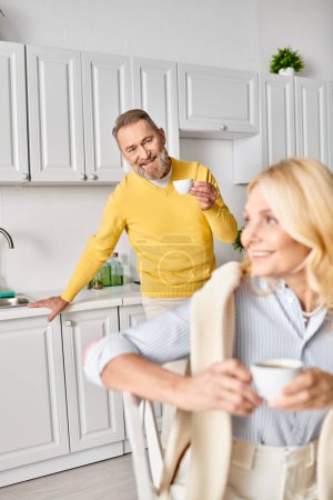 Una pareja madura y cariñosa en acogedora ropa de casa de pie juntos en una cocina en casa, compartiendo un momento especial.