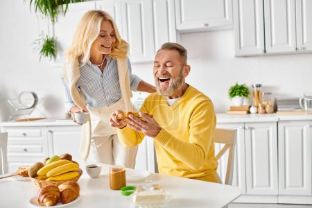 A mature man and woman dressed in cozy homewear sitting at a kitchen table, enjoying a moment together in their home.