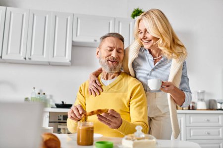 A mature man and woman in cozy home attire stand together in a warm kitchen, enjoying each others company.