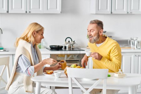 Una pareja madura y cariñosa en ropa de casa acogedora disfrutando de una comida juntos en una mesa en su cocina en casa.