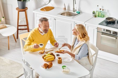 Ein reifer Mann und eine reife Frau genießen ein gemütliches Essen zusammen an einem Tisch in ihrer Küche und teilen Liebe und Lachen.