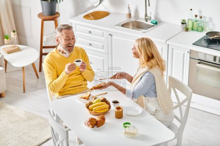 Un couple aimant mature dans des vêtements confortables assis ensemble à une table de cuisine, profitant d'un moment de convivialité à la maison.