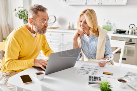 A mature man and woman in homewear sitting at a table, focused on a laptop screen.