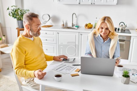 Téléchargez les photos : Un couple aimant mature dans des vêtements confortables assis ensemble à une table, travaillant sur un ordinateur portable. - en image libre de droit