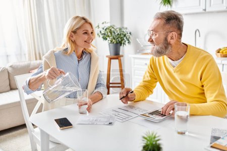 Ein reifer Mann und eine reife Frau in gemütlicher Hauskleidung sitzen an einem Tisch und konzentrieren sich darauf, einen Taschenrechner für finanzielle Berechnungen zu benutzen..
