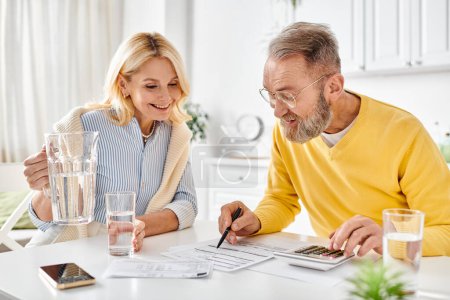 A mature man and woman in cozy homewear sit at a table, engaged in using a calculator together.