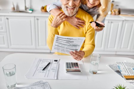 A mature man and woman in cozy homewear sit at a table working on a calculator, seemingly budgeting and planning together.