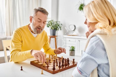 A mature, loving couple in cozy homewear engaged in a competitive game of chess, focused on the board before them.