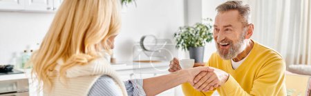 Photo for A mature man and woman in cozy homewear sit at the kitchen table, shaking hands in a gesture of agreement. - Royalty Free Image