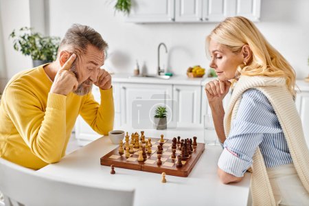 A mature loving couple in cozy homewear engaged in a strategic chess battle at a table in their kitchen at home.