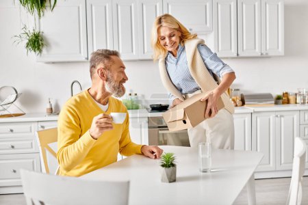 A mature man and woman in cozy homewear are seen moving boxes into a kitchen together at home.