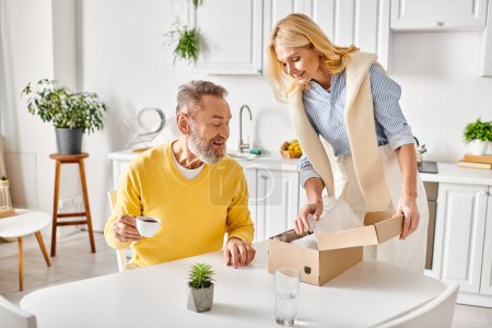 Photo for A mature loving couple in cozy homewear, opening a box to reveal a plant inside, sharing a moment of joy and discovery in their kitchen. - Royalty Free Image