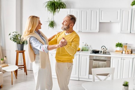 Ein reifes Liebespaar in kuscheliger Homewear tanzt zu Hause in der Küche fröhlich zusammen.