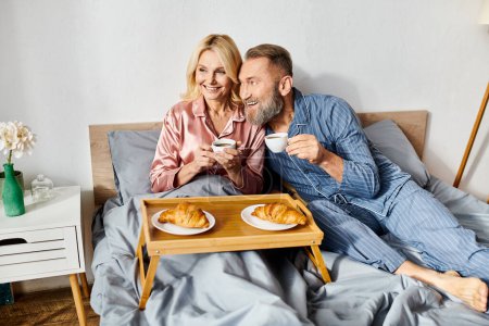 A mature loving couple in cozy homewear sitting on a bed, enjoying a tray of food together in their bedroom.