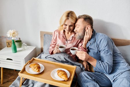 A mature loving couple in cozy homewear enjoying a relaxing morning on their bed with coffee and pastries.
