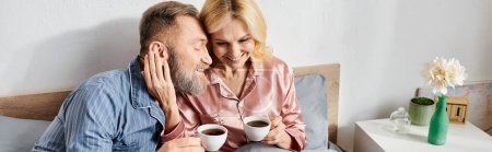 Téléchargez les photos : Un couple aimant mature, vêtu de vêtements confortables, s'asseoir sur un lit tenant des tasses de café, passer du temps de qualité ensemble. - en image libre de droit