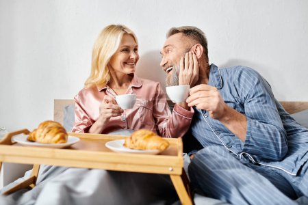 Foto de Una pareja amorosa madura en ropa de casa acogedora sentada en una cama, saboreando el café juntos en un entorno tranquilo dormitorio. - Imagen libre de derechos