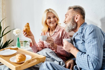 A mature loving couple in casual homewear enjoying a meal together at a table in their bedroom.