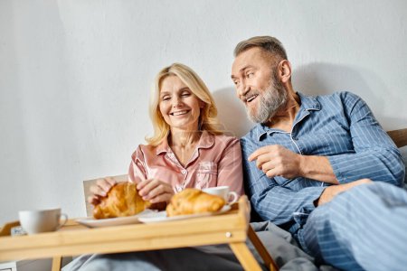 Photo for A mature loving couple in homewear sitting on a couch, enjoying a tray of croissants together in a cozy setting. - Royalty Free Image