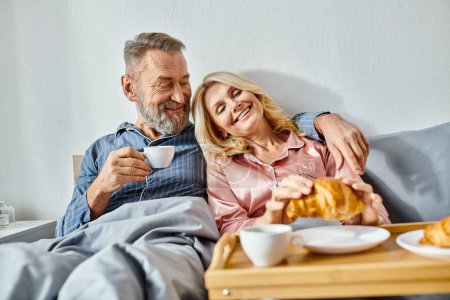 Téléchargez les photos : Couple d'âge mûr dans des vêtements confortables se détendre sur le canapé, partager un plateau de nourriture délicieuse. - en image libre de droit