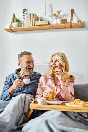 Ein erwachsener Mann und eine erwachsene Frau genießen eine Mahlzeit, während sie auf einer bequemen Couch in ihrem gemütlichen Schlafzimmer sitzen und in Homewear gekleidet sind.