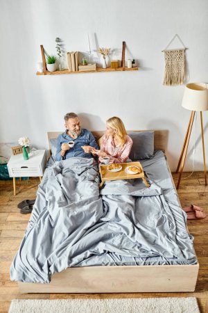 A mature loving couple in cozy homewear, sitting closely together on a bed, sharing a peaceful and tender moment.