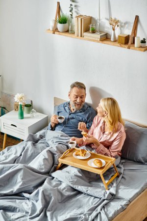 A mature man and woman in cozy homewear sitting together on a bed, sharing a quiet moment of togetherness.