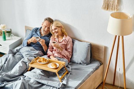 Mature loving couple in cozy homewear sitting on bed, enjoying each other