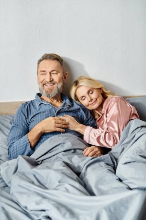 A mature man and woman cuddling in bed, wearing cozy homewear, enjoying each others company in their bedroom.
