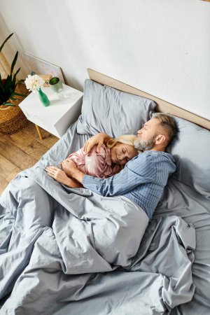 A mature loving couple in cozy homewear laying together in bed, facing each other with serene expressions.
