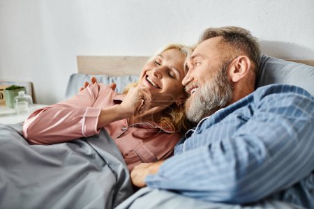 A mature man and woman in cozy homewear laying together in bed, sharing a peaceful moment of intimacy and connection.