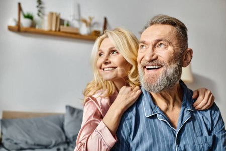 A mature man and woman in cozy homewear are smiling together in their bedroom, radiating love and happiness.