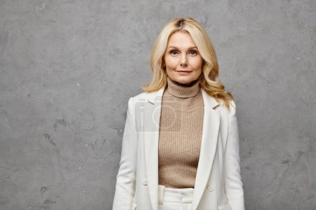 A mature woman exudes elegance in a fashionable white jacket and turtle neck sweater against a gray backdrop.