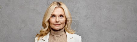 Mature woman exudes sophistication in white jacket and turtle neck sweater against a gray backdrop.