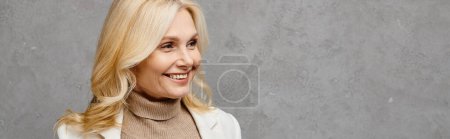 Téléchargez les photos : Élégante femme mature aux cheveux blonds et à la veste blanche souriant avec charme sur fond gris. - en image libre de droit