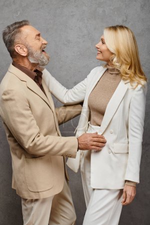 Téléchargez les photos : Un homme et une femme mûrs en tenue debonair se tiennent côte à côte, exsudant l'amour et la grâce sur un fond gris. - en image libre de droit