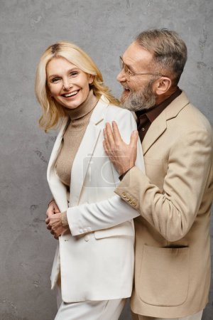 Téléchargez les photos : Couple d'amour mature en tenue debonair se tiennent gracieusement ensemble sur un fond gris. - en image libre de droit