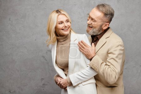 Téléchargez les photos : Elégant, homme et femme mûrs en tenue debonair debout ensemble, exsudant l'amour et la sophistication, dans un contexte gris. - en image libre de droit