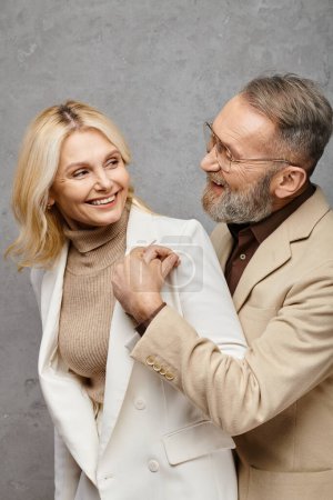 A elegant, mature man helps a woman put on her coat in a debonair pose against a gray backdrop.
