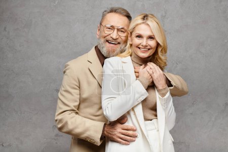 Un hombre y una mujer maduros, exudando elegancia, posan juntos con un atuendo elegante sobre un fondo gris.