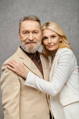 Mature couple dressed elegantly pose together in front of a gray backdrop.