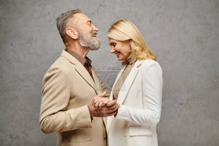 Mature, elegant couple in debonair attire embrace, holding hands lovingly against a gray backdrop.