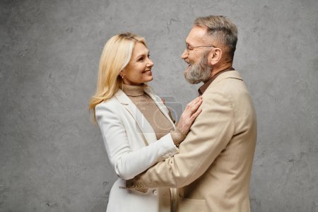 Eleganter, reifer Mann und Frau in stilvoller Kleidung posieren gemeinsam vor grauem Hintergrund.