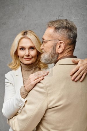 A man and a woman in elegant attire embracing each other affectionately against a gray backdrop.