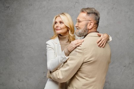 Ein reifer Mann und eine reife Frau, elegant gekleidet, umarmen sich zärtlich vor grauem Hintergrund.