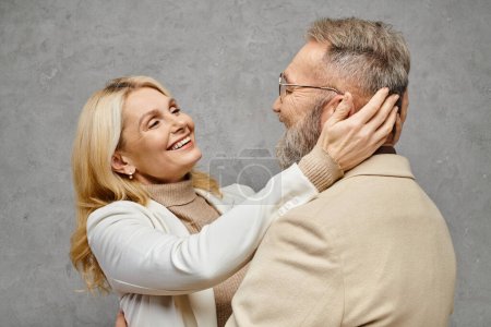 A mature man and woman in elegant attire embrace lovingly against a gray backdrop.
